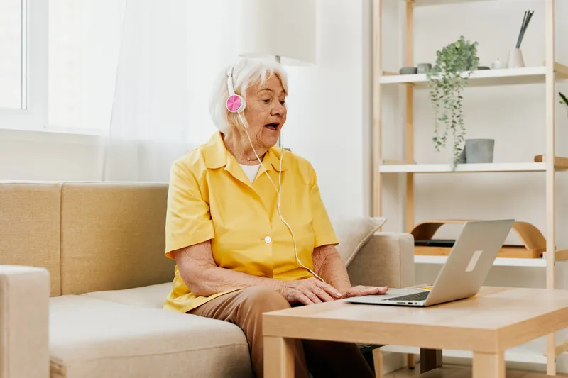 Пожилая женщина в наушниках сидит дома на диване с ноутбуком и работает в жёлтой рубашке перед окном, на её лице удивление