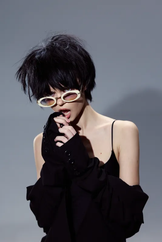 Stylish woman with short black hair wearing trendy sunglasses and black outfit posing on gray background.
