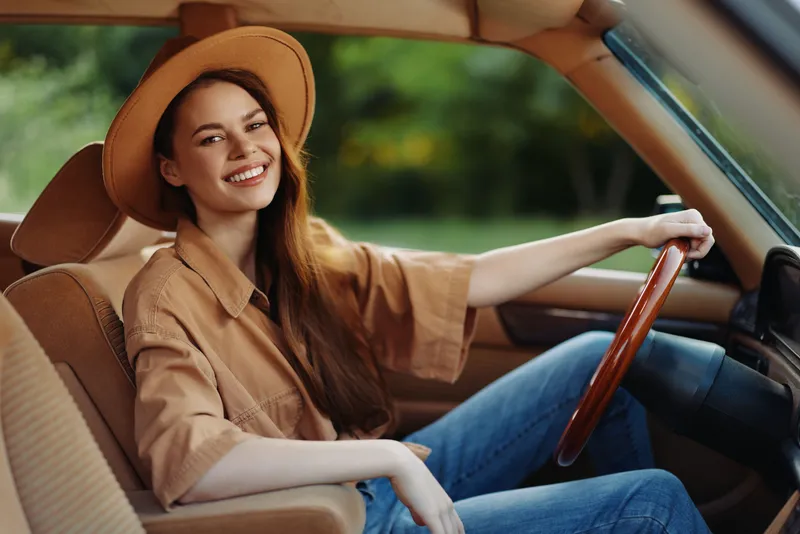 Happy young woman with long hair smiling in vintage car on a sunny day, wearing a brown outfit and hat, surrounded by green nature.
