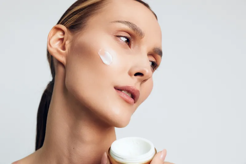 Young woman closeup applies facial cream on face with fingers holding skincare jar, fresh moisturizer on cheek, beauty spa routine, clean studio background, skin care concept, copy space