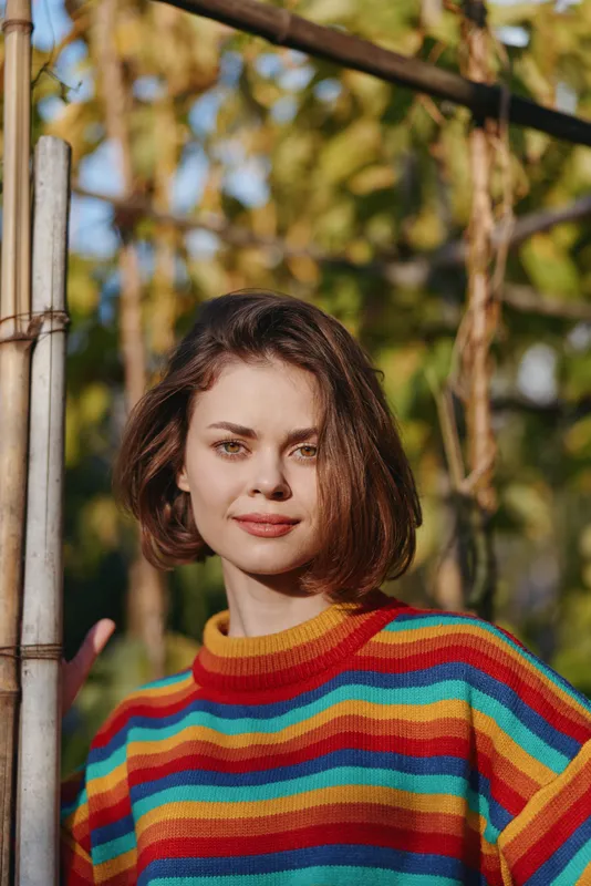 Woman portrait in colorful sweater smiling outdoors with short hair and warm sunlight and garden background, casual fashion portrait of a confident young adult enjoying natural light.