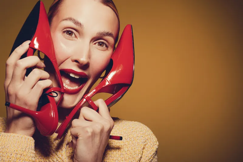 Happy woman with red high heels holds shoes near face, smiling with open mouth. Yellow background and warm lighting create joyful and fashionable mood for style and beauty concept.