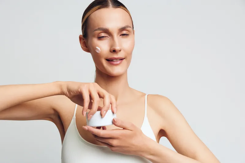 A young woman in a white tank top applies moisturizer to her face with a small amount on her cheek. She holds a white jar of cream in a bright studio setting. Concept for skincare routine.