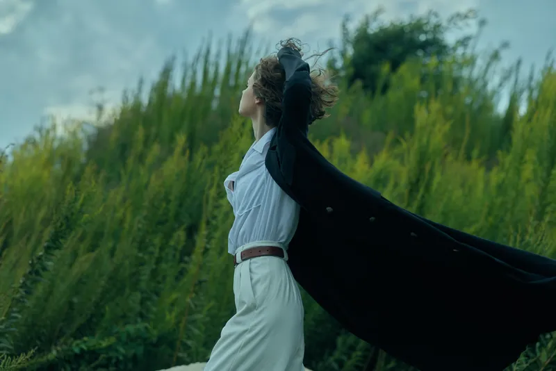 Cape billows behind a runner in white as wind sweeps through tall grass, capturing freedom, motion, and outdoor fashion in a bright expansive landscape