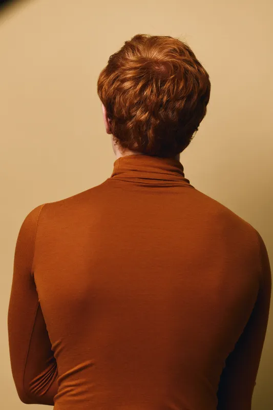 Man with red hair wearing brown turtleneck sweater facing back on neutral background with soft lighting and minimalistic style portrait photograph