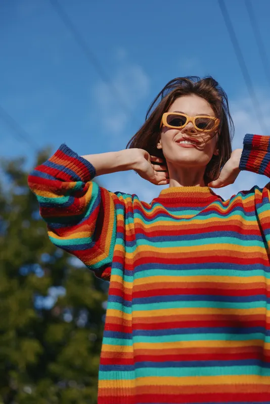 Young woman in a colorful striped sweater and yellow sunglasses smiling outdoors under a bright blue sky, hands behind neck, casual fashion lifestyle portrait.