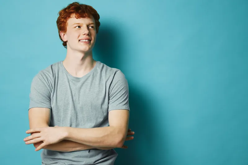 Young man with red hair in gray t shirt standing with arms crossed looking sideways against blue background for casual style confidence and youth portrait.