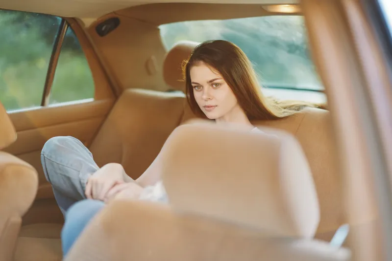 Young woman with long brown hair, wearing casual clothing, sitting in a car, appearing contemplative and serene, with a blurred background of nature.