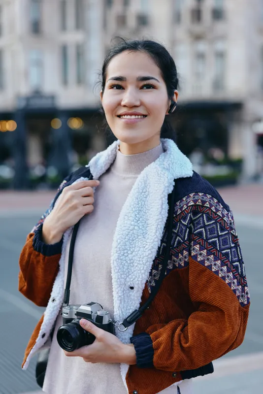 Woman photographer outdoors with camera, wearing a cozy jacket, urban street backdrop, exploring city scenes and ready to capture moments.