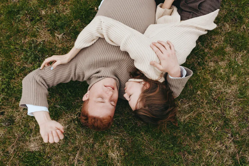 A couple rests on a grassy field in cozy knitwear, sharing a quiet moment outdoors, hands linked, eyes closed. A lifestyle scene that captures comfort, connection, and simple joy in nature.