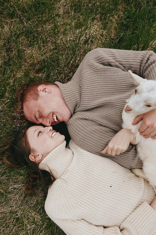 Smiling couple lying on grass with a playful goat kid nearby, embracing an outdoors lifestyle. Warm light and soft textures convey relaxation, happiness, and natural harmony.