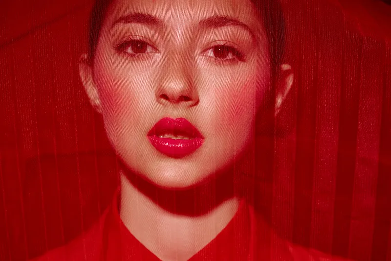 Close-up portrait of a woman with vibrant red lipstick and makeup, textured red veil creating dramatic fashion beauty concept with artistic lighting and striking expression.