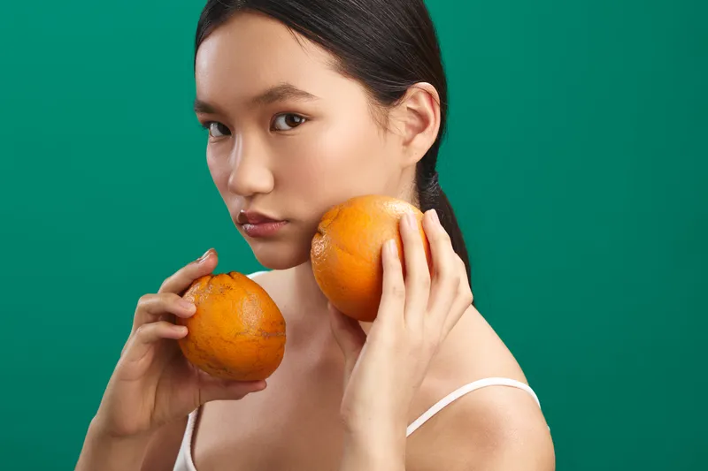 asian woman presents a calm expression while holding two oranges in a bright studio with a green backdrop, creating a fresh, colorful lifestyle image focused on fruit.
