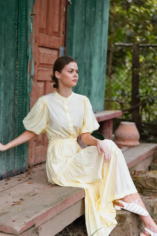 Woman in a pale yellow dress sits outdoors on steps by a green doorway, calm and elegant, capturing a serene daytime mood in a lush garden.
