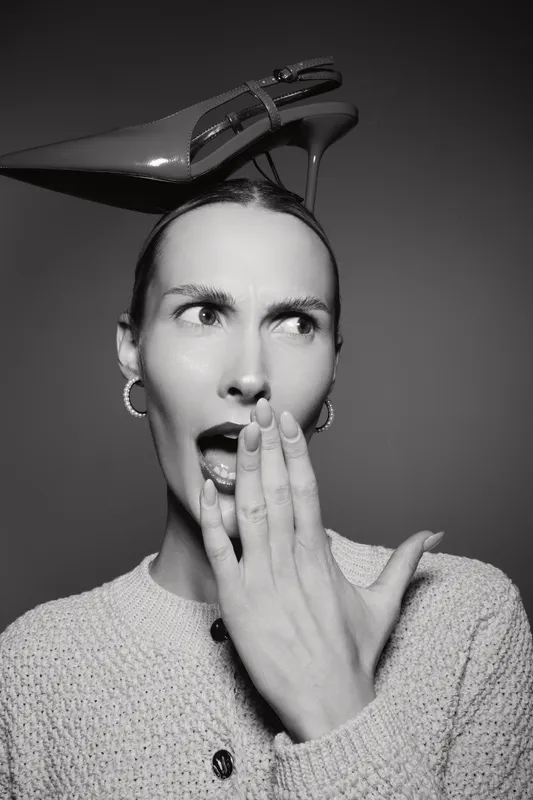 Young caucasian woman with light skin and short hair covers her mouth in surprise, balancing a high heel shoe on her head in studio.