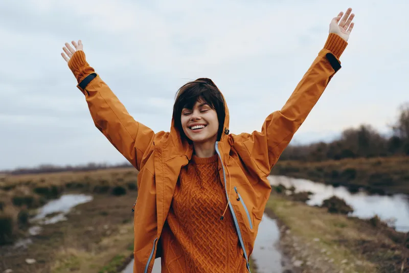 Woman outdoors on rural path smiling, arms raised, wearing orange jacket and sweater, cheerful lifestyle moment, nature scenery, countryside fields, cloudy sky, natural light