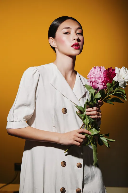 Fashion model with glossy sleek hair and bold red lipstick poses holding fresh vibrant flowers on orange background. Artistic beauty concept with natural elegance light.