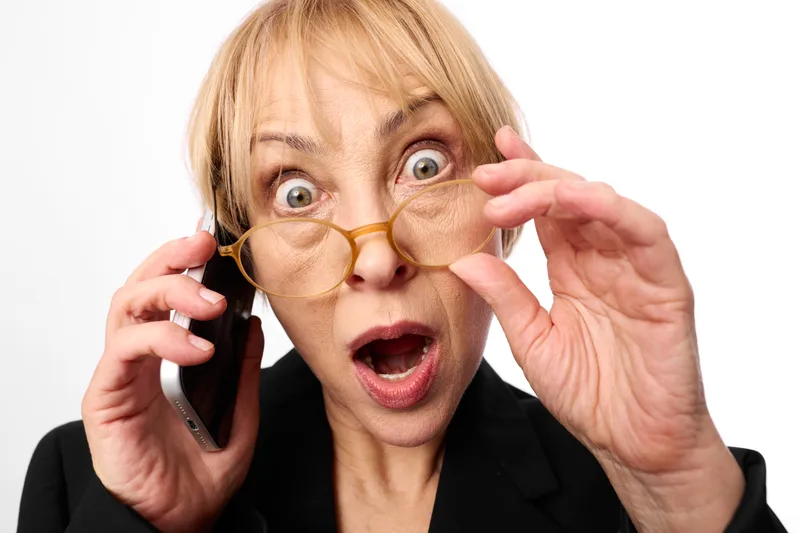 Surprised woman with glasses answers a phone call, eyes wide with shock and stress, holding smartphone near ear, concept for communication panic