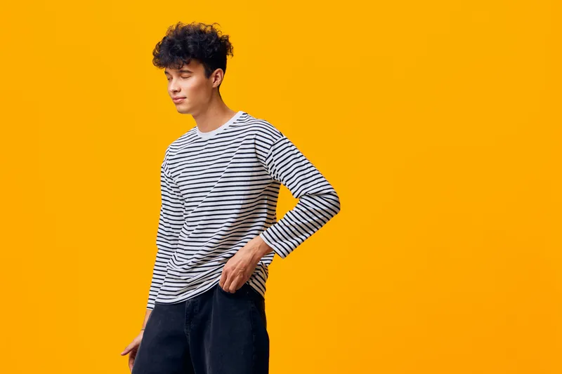 young man with curly hair wearing striped shirt and black pants posing thoughtfully against vibrant orange background, casual style, modern fashion, studio shot