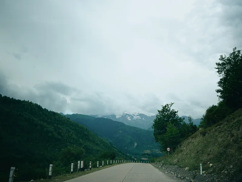 Mountain road through a valley with forested hills, cloudy sky, guardrails, and open asphalt offering a remote scenic drive, rugged terrain, and calm nature in a wide alpine landscape