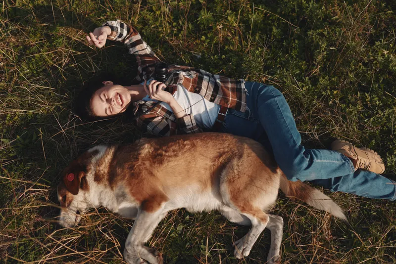 Woman lying on grass beside a friendly dog outdoors, relaxed moment in nature with a plaid shirt, casual stroll memory, and calm companionship under open sky.