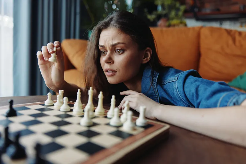 Женщина играет в шахматы перед диваном, на столе в гостиной разложены шахматные фигуры