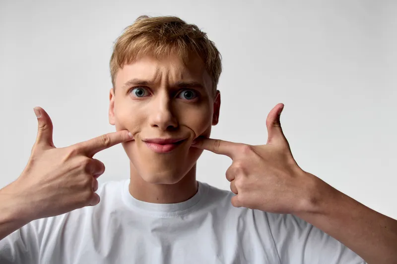Playful face, funny expression, hands pulling lips, young man, silly pose, facial expression in a casual studio shot with closeup lighting and cheeky humor.
