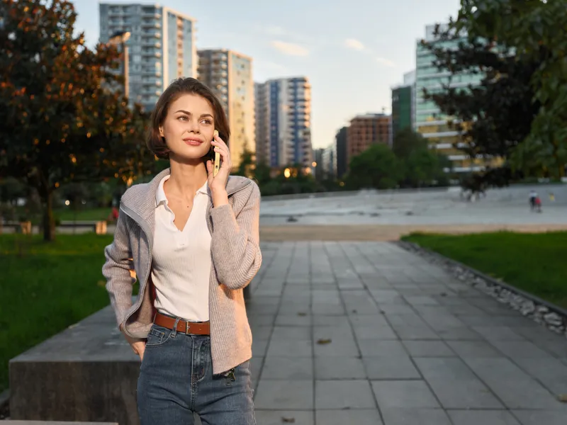 A confident woman in smart casual attire stands in an urban park, talking on a smartphone. She smiles subtly, with modern buildings in the background, conveying a dynamic, professional atmosphere.