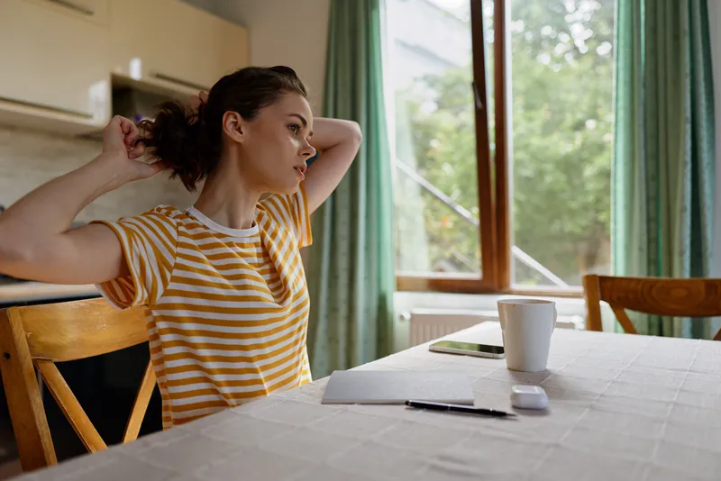 A young woman with a striped t shirt ties her hair back at a sunlit kitchen table, capturing a candid morning routine moment and quiet focus before starting her day, feeling organized.