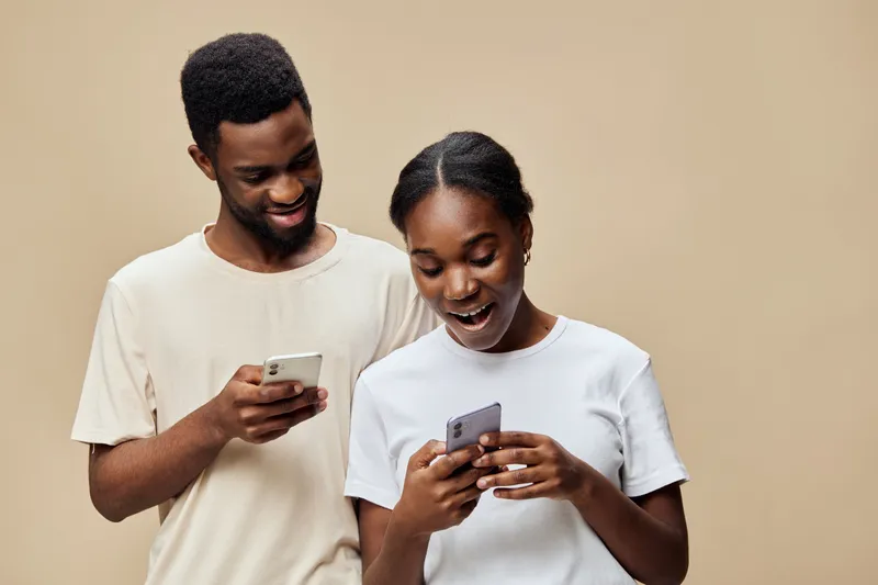 Young couple enjoying time together, smiling while using smartphones against a warm beige background, showcasing modern lifestyle and connection