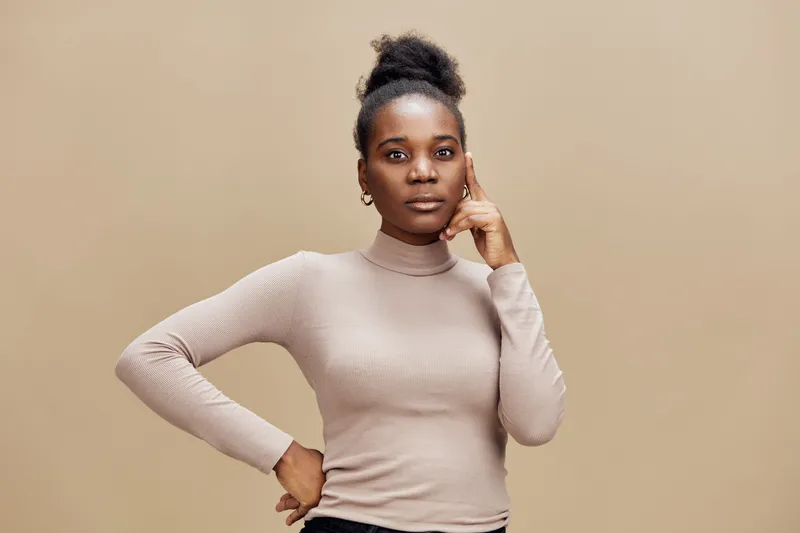 Young African woman in a beige turtleneck top, thoughtfully posing against a soft brown background, embodying confidence and contemplation in a minimalist style