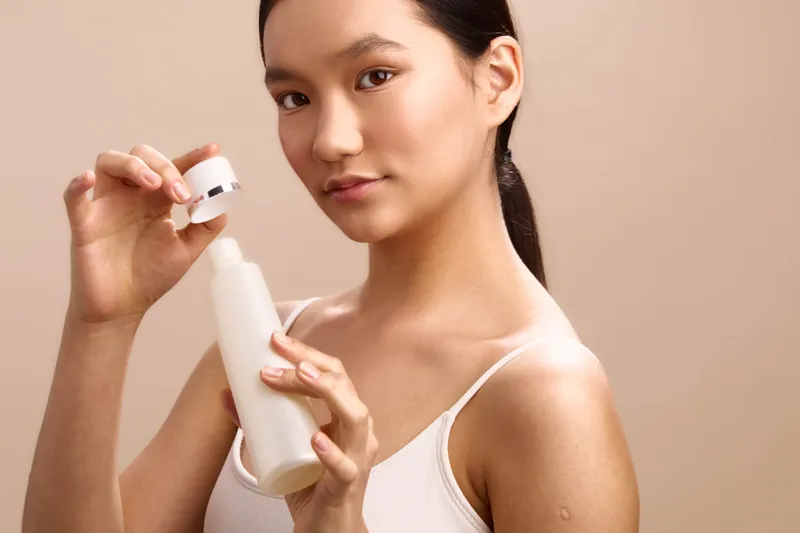 Asian woman demonstrates skincare routine, holding a small jar and a white bottle. She smiles softly, projecting calm confidence and a focus on personal wellness.