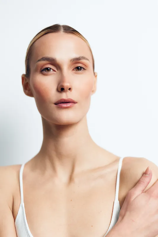 Close up portrait of calm young woman with natural skin and pulled back hair wearing white tank top isolated on white background with soft lighting and gentle expression