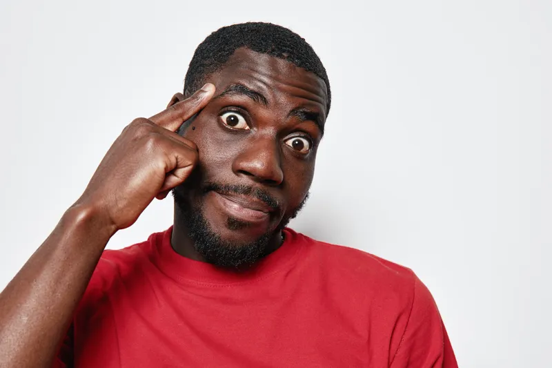 African American man with dark skin wearing red shirt pointing to head with finger, thinking and showing expression of idea or realization on white background.