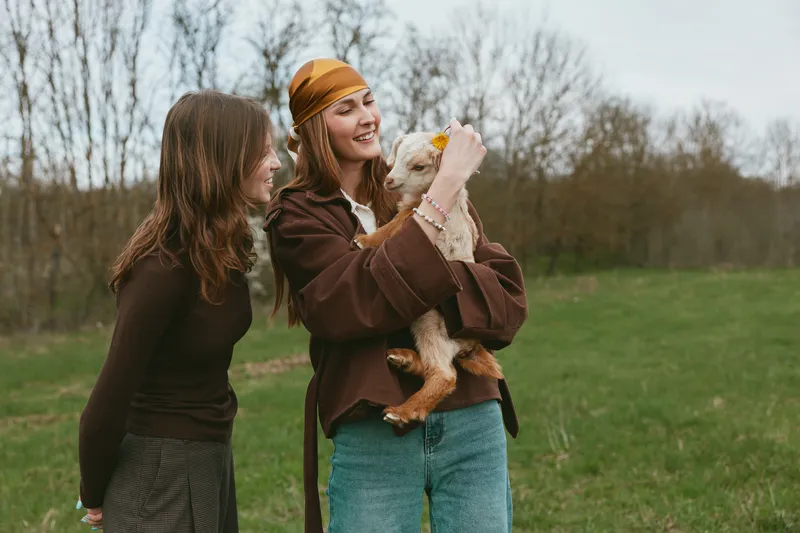 Lifestyle scene with two women and a curious goat kid outdoors, showcasing friendly affection, playful interaction and natural smiles in a field setting under a clear sky, rural atmosphere and casual
