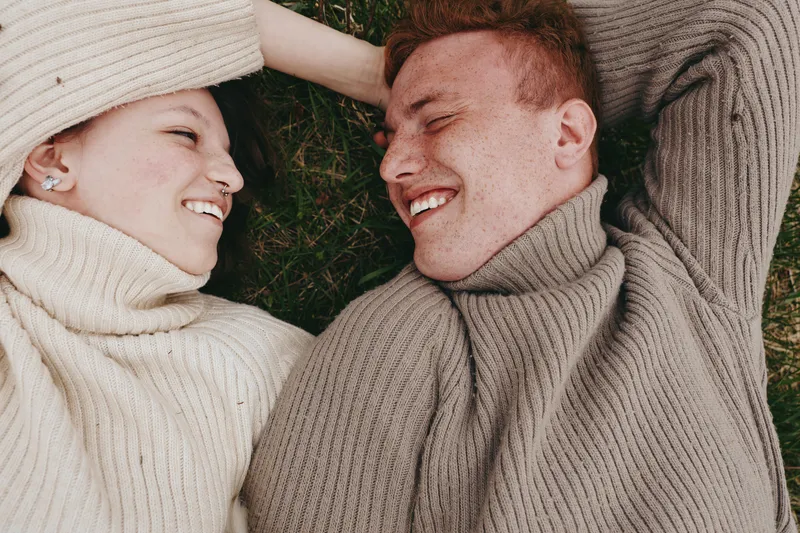 Joyful couple lying on grass, smiling at each other in cozy sweaters, outdoor moment with soft breeze and sunlit glow, showcasing relaxed lifestyle and intimate connection