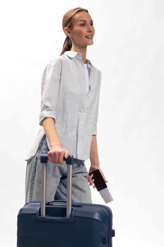 Young woman in casual clothes pulls blue suitcase with one hand and holds passport in the other. Travel concept with happy female traveler against white background.