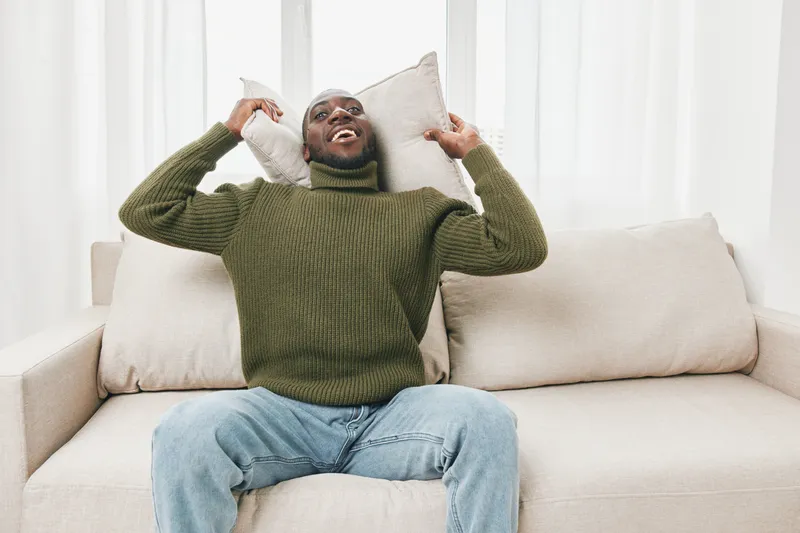 Happy Black man relaxing on a cozy sofa with pillows, wearing a green sweater, enjoying a peaceful moment indoors, conveying comfort and joy