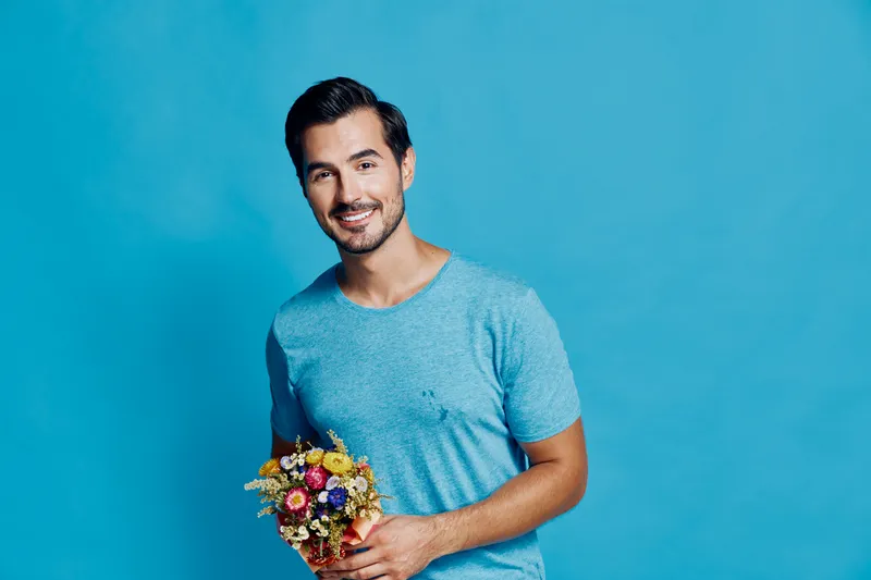 Cheerful smiling young man holding a colorful bouquet of flowers, wearing a casual blue t-shirt, standing against a vibrant blue background. People lifestyle concept.