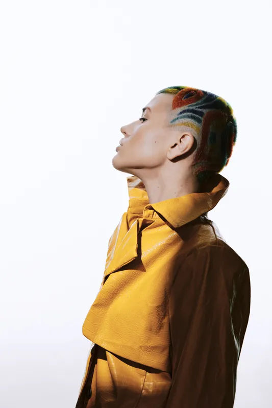 Woman with colorful patterned shaved hair wearing a mustard yellow jacket standing in profile with a bright minimalist background, showcasing modern fashion and unique style.