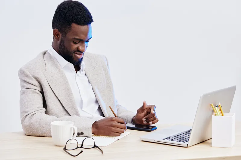 Professional young African American man working at desk with laptop, using smartphone, taking notes, dressed in beige blazer and white shirt, modern office environment with white background. Business 