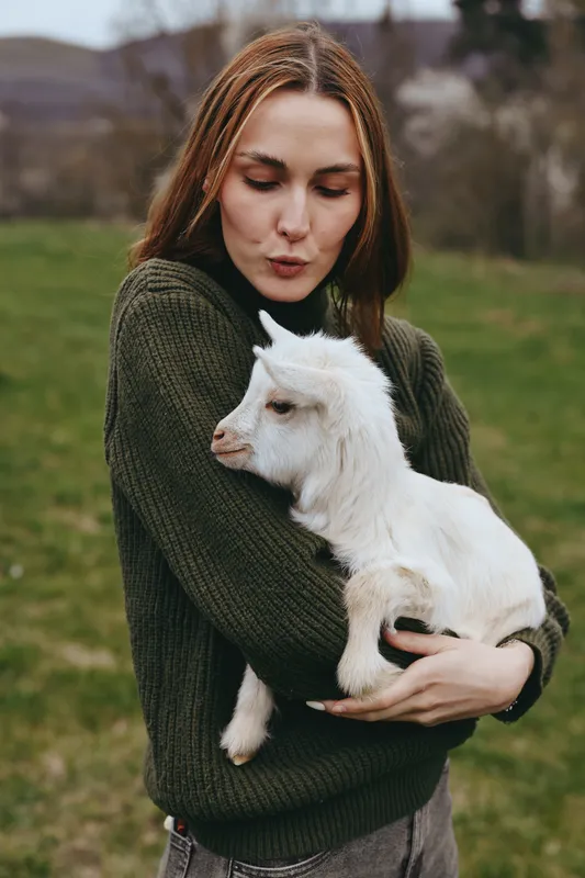 A woman in a dark sweater holds a young goat kid close in a sunny field, showing gentle affection and a strong bond with animals. Lifestyle moment outdoors, celebrating nature and rural living.