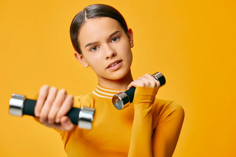 Young woman fitness portrait holding two chrome dumbbells, wearing mustard long sleeve top, focused gaze and poised posture against a vibrant orange studio background, active lifestyle.