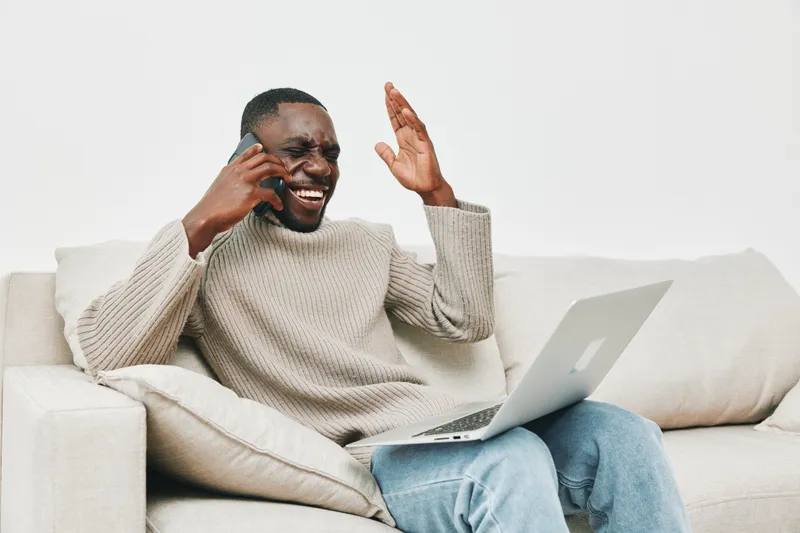 Happy young Black man cheerfully talking on the phone while sitting on a sofa with a laptop, showcasing joy and productivity in a modern living space