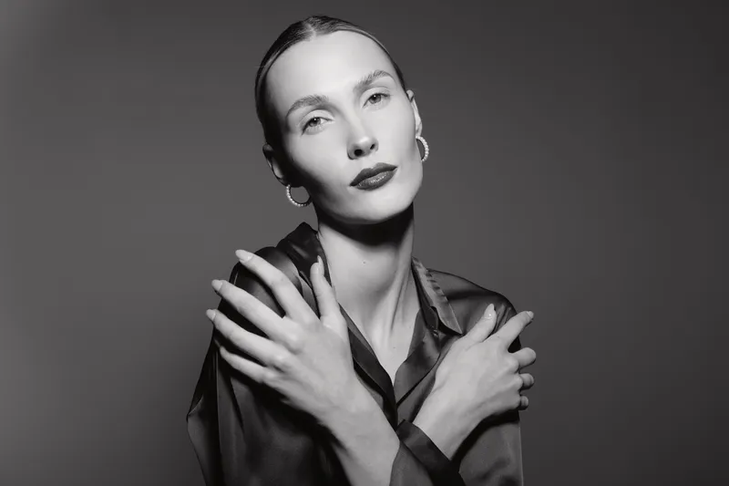 Portrait of a pale white woman with slicked back hair and hoop earrings poses with crossed arms in a dark satin blouse against a plain grey background. Concept of elegance and minimalism.