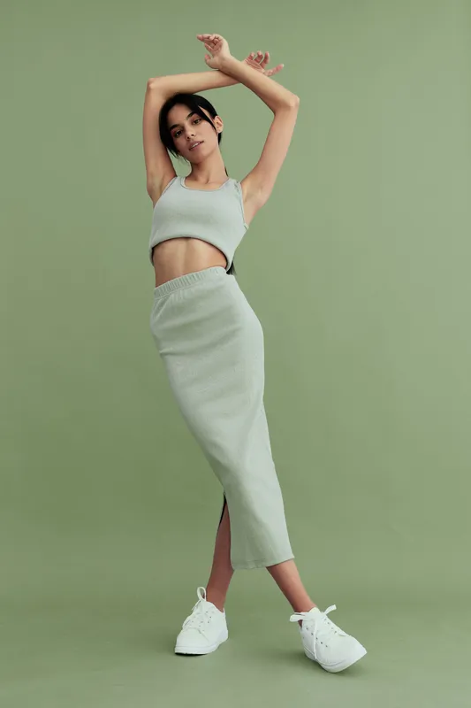 Young woman posing gracefully in a stylish ribbed crop top and matching skirt set with white sneakers against a soft green background in a contemporary studio.