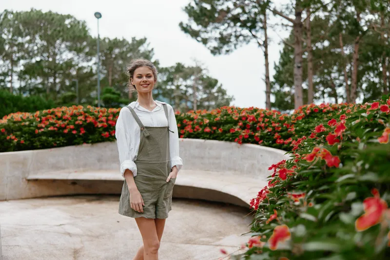 Young woman smiling in a garden with flowers, wearing stylish green overalls and a white shirt, conveying happiness in a bright outdoor setting