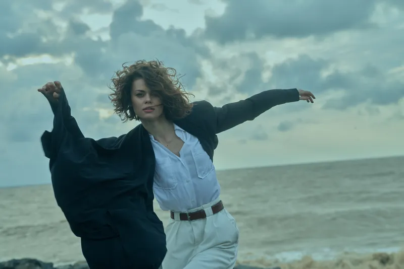 Woman on a windy beach in a white shirt and dark jacket, arms outstretched, embracing freedom and motion against a cloudy sea backdrop.