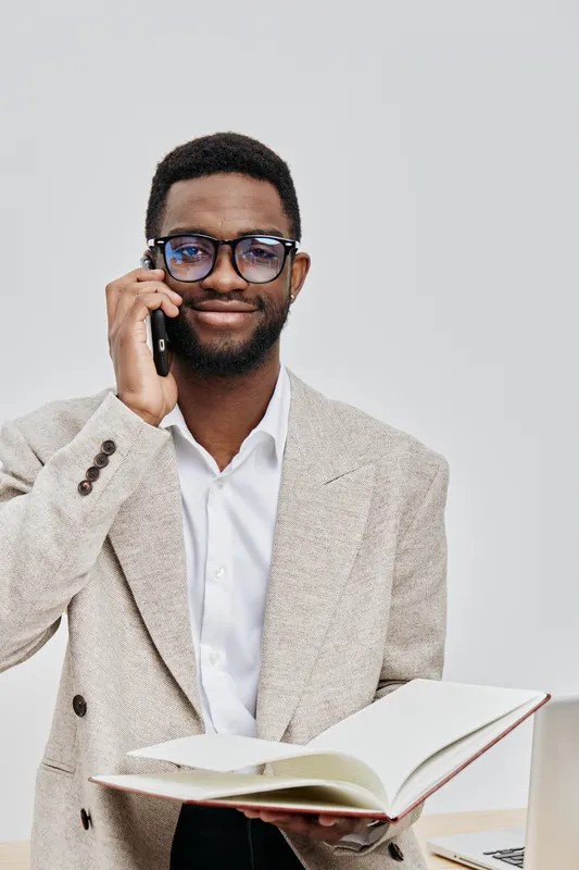 Confident young man using mobile phone while holding an open book in a professional setting, wearing glasses and a beige blazer, demonstrating communication and knowledge sharing, studio portrait. Peo