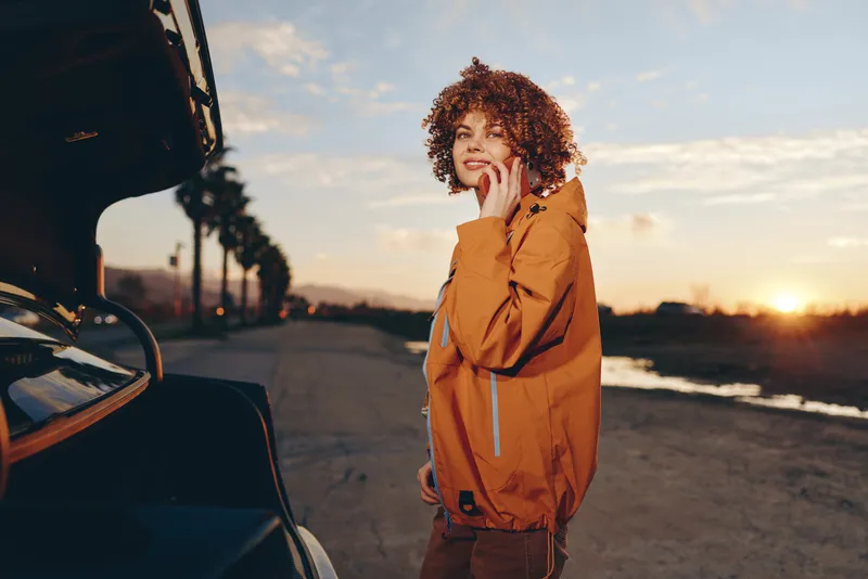 woman lifestyle sunset car jacket travel outdoor road curly hair, young adult stands near parked car on scenic roadway at golden evening, orange jacket, natural smile, wind, backlight portrait
